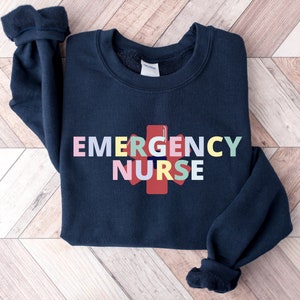 Puede incluir: Una sudadera azul marino con las palabras "EMERGENCY NURSE" en letras de colores y un s&iacute;mbolo m&eacute;dico rojo.