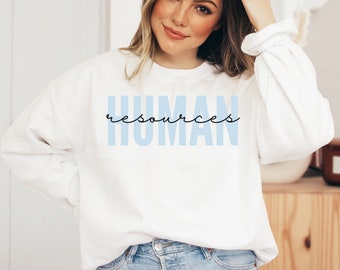 May include: White sweatshirt with the words "HUMAN resources" printed in light blue and black. The sweatshirt has long sleeves and a crew neck. The text is in a sans-serif font for "HUMAN" and a script font for "resources".