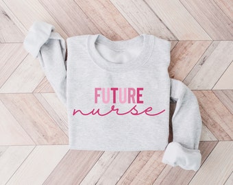 May include: A light gray sweatshirt with the words "FUTURE nurse" printed in shades of pink. The sleeves are rolled up, and the sweatshirt is displayed on a light brown wooden surface.