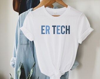 May include: A white t-shirt with the text "ER TECH" in shades of blue. The shirt is hanging on a wooden hanger with other clothing items, including a denim shirt and a brown hat. The shirt is made of a soft material.