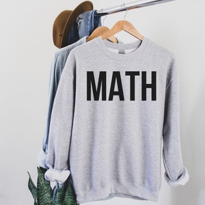 May include: A light gray sweatshirt with the word "MATH" printed in large, black capital letters. The sweatshirt is hanging on a wooden hanger. A denim shirt and a brown hat are also hanging on the rack.