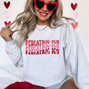 Op de afbeelding: Een grijs sweatshirt met de tekst "PEDIATRIC ICU" drie keer herhaald in een retro-lettertype met roze harten.