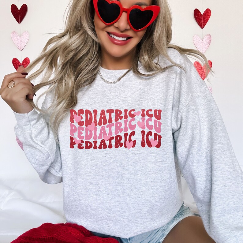 Op de afbeelding: Een grijs sweatshirt met de tekst "PEDIATRIC ICU" drie keer herhaald in een retro-lettertype met roze harten.