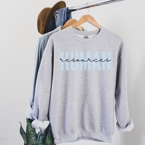 May include: A light gray sweatshirt with the words "HUMAN resources" printed on the front in light blue and black script. The sweatshirt is hanging on a wooden hanger with other clothing items.