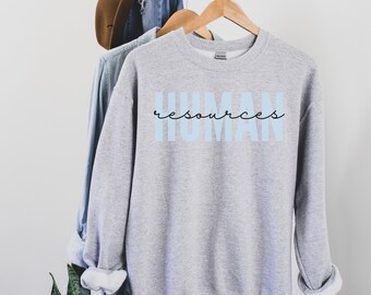 May include: A light gray sweatshirt with the words "HUMAN resources" printed on the front in light blue and black script. The sweatshirt is hanging on a wooden hanger with other clothing items.