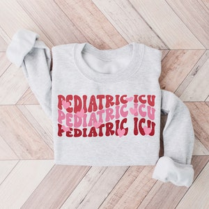 Op de afbeelding: Een grijs sweatshirt met de tekst "PEDIATRIC ICU" drie keer herhaald in een retro-lettertype met roze hartjes.
