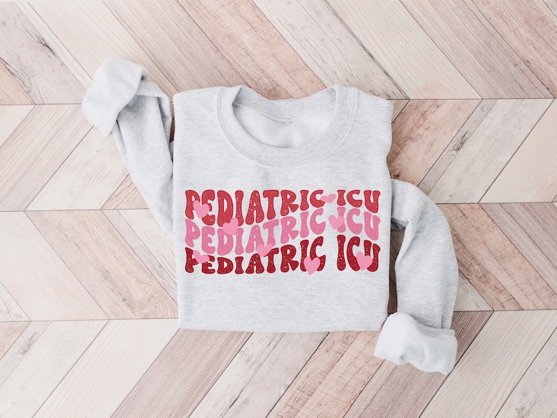 Op de afbeelding: Een grijs sweatshirt met de tekst "PEDIATRIC ICU" drie keer herhaald in een retro-lettertype met roze hartjes.