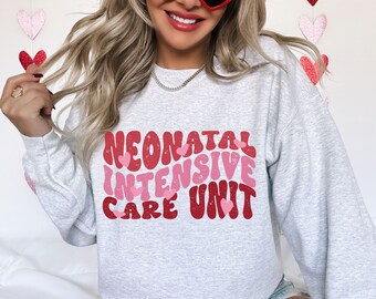 May include: A light gray sweatshirt with the text "NEONATAL INTENSIVE CARE UNIT" in red and pink lettering. The design includes heart shapes. The sweatshirt is a casual style.