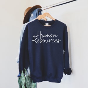 May include: Navy blue sweatshirt with the words "Human Resources" in white script. The sweatshirt is hanging on a wooden hanger, with a denim shirt and a brown hat in the background.