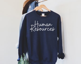 May include: Navy blue sweatshirt with the words "Human Resources" in white script. The sweatshirt is hanging on a wooden hanger, with a denim shirt and a brown hat in the background.