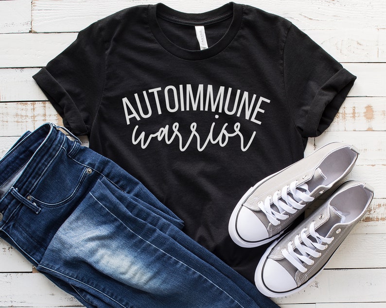 May include: Black t-shirt with white text that reads "AUTOIMMUNE warrior". The shirt is paired with blue jeans and gray sneakers.