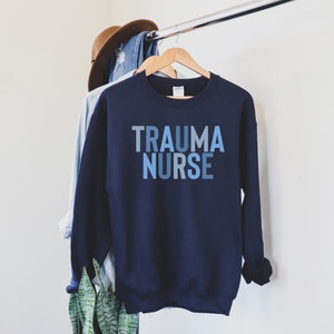 May include: A navy blue sweatshirt with the text "TRAUMA NURSE" printed in blue and gray ombre lettering.