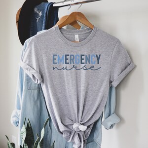 May include: A heather gray t-shirt with the words "EMERGENCY nurse" printed in blue. The shirt is hanging on a wooden hanger with a blue denim shirt and a brown hat in the background.