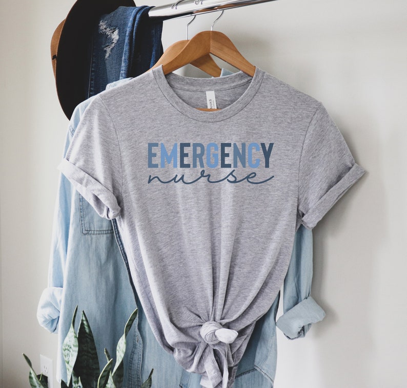 May include: A heather gray t-shirt with the words "EMERGENCY nurse" printed in blue. The shirt is hanging on a wooden hanger with a blue denim shirt and a brown hat in the background.