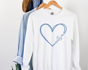 May include: White sweatshirt with a blue heart design and the word "autism" written inside the heart. The sweatshirt is hanging on a wooden hanger. A denim shirt and a brown hat are visible in the background.