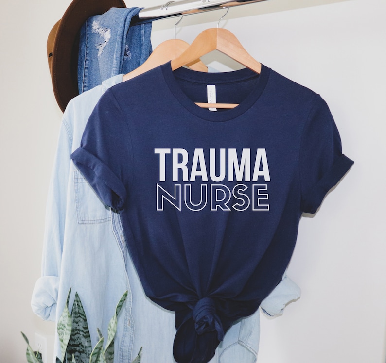 May include: A navy blue t-shirt with white text that reads "TRAUMA NURSE". The shirt is folded and hanging on a wooden hanger.