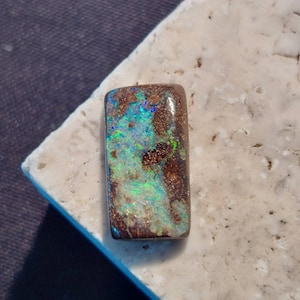 May include: A rectangular, brown opal gemstone with green, blue, and white iridescent flashes. The opal is set on a white, textured surface.