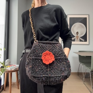 May include: A handmade black crochet bag with a gold chain strap and a bright orange flower applique. The bag is made of a textured yarn with flecks of color throughout. The bag is a shoulder bag style.