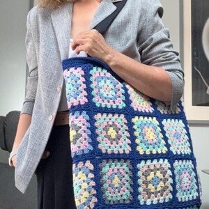 May include: A handmade, blue crochet tote bag with a colorful granny square pattern. The bag features squares in pastel shades of pink, yellow, and blue, creating a vibrant and unique design. The bag has a black strap.