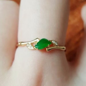 May include: A gold ring featuring a green jade stone set within a branch design. The ring is worn on a finger.