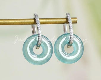 May include: A pair of jade earrings with a circular, donut-shaped design. The earrings are a light teal color and are suspended from silver hoops encrusted with small, clear gemstones. The earrings are displayed on a gold-colored bar against a soft, blurred background.