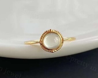 May include: A gold ring with a white stone set in a decorative bezel. The ring is on a white surface.