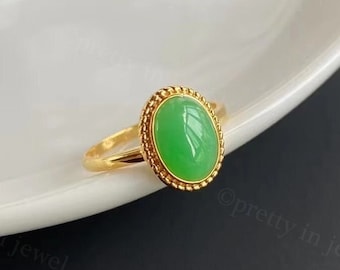 May include: A gold ring featuring a vibrant green oval gemstone. The gemstone is set within a beaded border, and the ring band is a simple, polished gold. The ring is displayed on a white, curved surface.