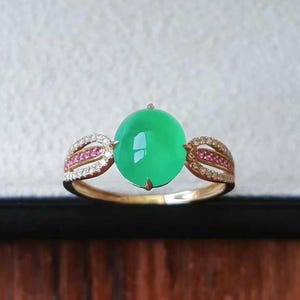 May include: A gold ring featuring a large, vibrant green jade cabochon as the centerpiece. The ring is accented with small pink and white gemstones. The words "Jadeite Jade Jewelry" are at the bottom of the image.
