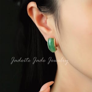 Pu&ograve; includere: Primo piano di un orecchino di giada con un bordo color oro. L'orecchino ha una forma curva e un colore verde vibrante. Il testo "Jadeite Jade Jewelry" &egrave; scritto in corsivo.