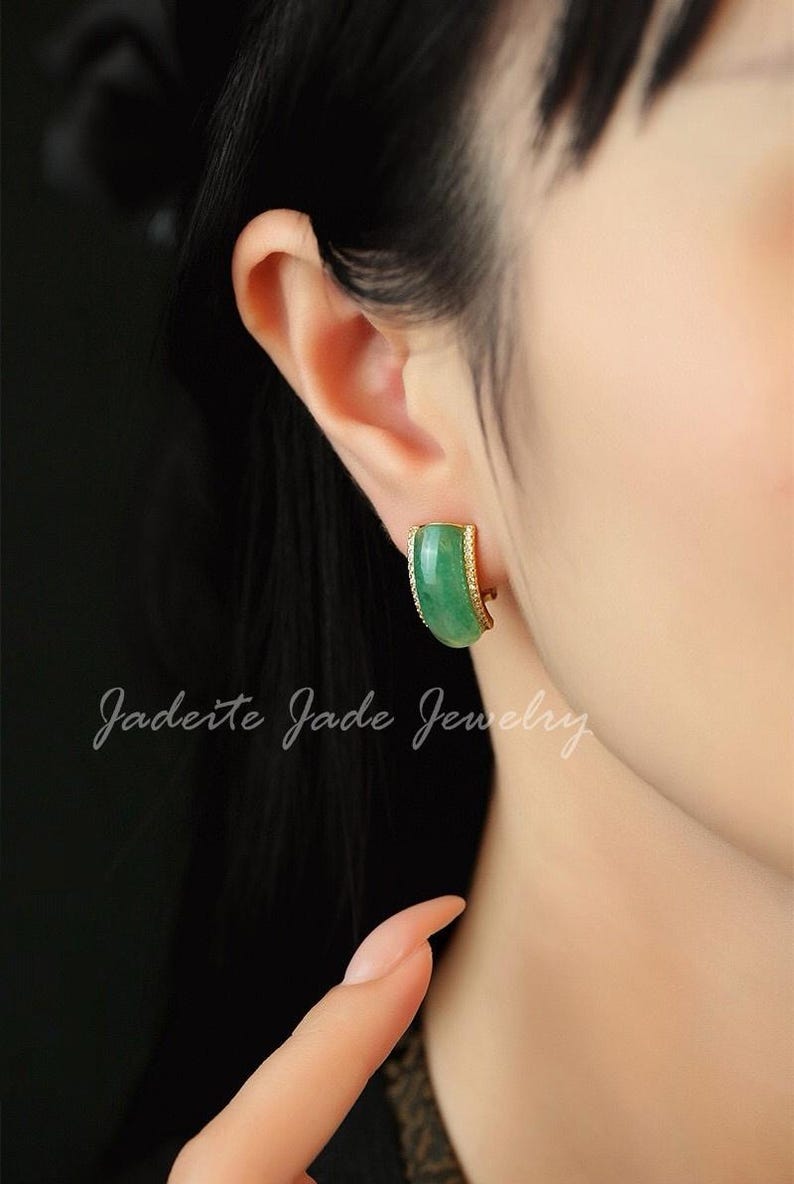 Pu&ograve; includere: Primo piano di un orecchino di giada con un bordo color oro. L'orecchino ha una forma curva e un colore verde vibrante. Il testo "Jadeite Jade Jewelry" &egrave; scritto in corsivo.