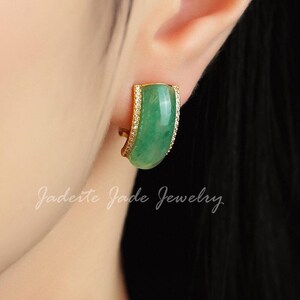 Pu&ograve; includere: Un orecchino dorato con una pietra di giada verde curva e rettangolare. L'orecchino &egrave; impreziosito da piccole pietre trasparenti lungo il bordo. Il testo "Jadeite Jade Jewelry" &egrave; scritto in corsivo.