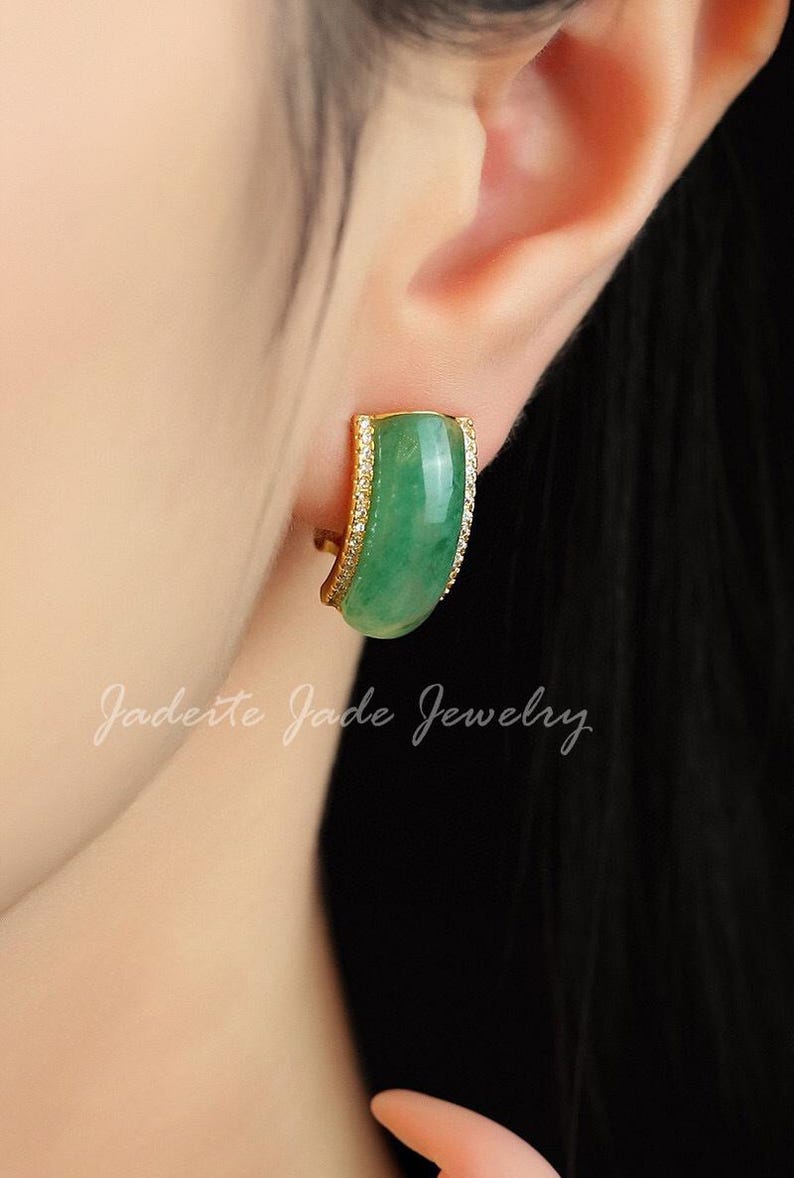 Pu&ograve; includere: Un orecchino dorato con una pietra di giada verde curva e rettangolare. L'orecchino &egrave; impreziosito da piccole pietre trasparenti lungo il bordo. Il testo "Jadeite Jade Jewelry" &egrave; scritto in corsivo.