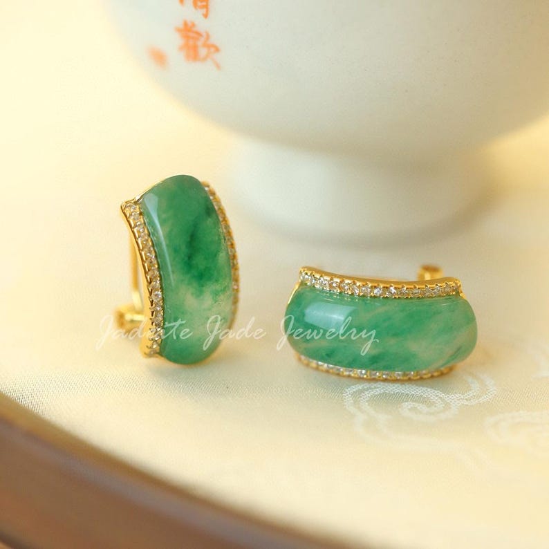 Pu&ograve; includere: Un paio di orecchini di giada. Ogni orecchino presenta una pietra di giada verde chiaro curva, incastonata in una montatura color oro. La montatura &egrave; impreziosita da piccole pietre trasparenti. Gli orecchini sono esposti su una superficie chiara.