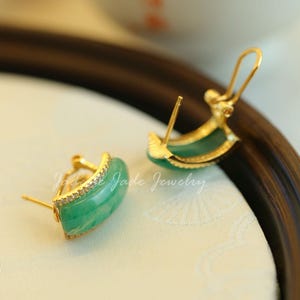 Pu&ograve; includere: Un paio di orecchini color oro con una pietra verde giada curva. Gli orecchini hanno una piccola fila di pietre trasparenti lungo il bordo superiore. Gli orecchini sono su una superficie color crema. Il testo "Jade Jewelry" &egrave; visibile.