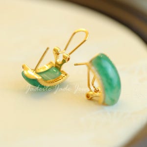 Pu&ograve; includere: Un paio di orecchini color oro con una pietra di giada verde curva. Gli orecchini hanno una chiusura a gancio e sono incastonati in un metallo color oro. Il testo "Jaderite Jade Jew" &egrave; visibile.