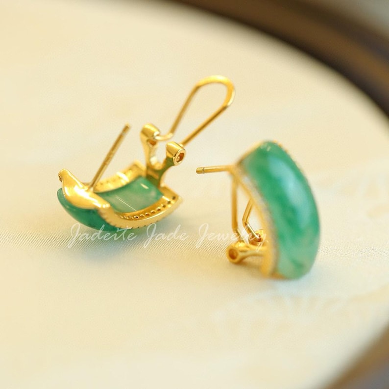 Pu&ograve; includere: Un paio di orecchini color oro con una pietra di giada verde curva. Gli orecchini hanno una chiusura a gancio e sono incastonati in un metallo color oro. Il testo "Jaderite Jade Jew" &egrave; visibile.