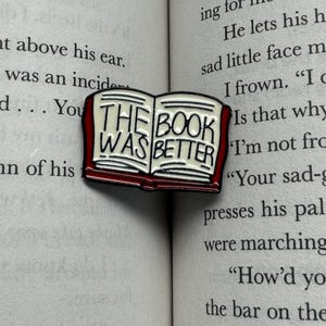 May include: A metal enamel pin shaped like an open book with a dark red cover and cream-colored pages. The text THE BOOK WAS BETTER is printed in black capital letters across the open pages of the pin, which rests on a printed book page.