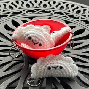 May include: Three white crocheted dumplings with black eyes and a smiling mouth. The dumplings are in a red bowl and have keychains attached to them.
