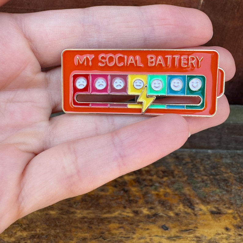 May include: A rectangular enamel pin with a red border and the text "MY SOCIAL BATTERY". The pin features a battery level indicator with sad to happy face emojis. A yellow lightning bolt graphic is in the center.