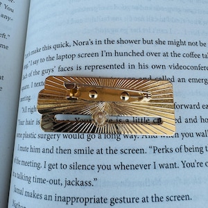 May include: A gold-colored metal pin with a sunburst design. The pin has a small, clear, round gem and the text "Your hair is a little cr". The pin is resting on the open pages of a book.