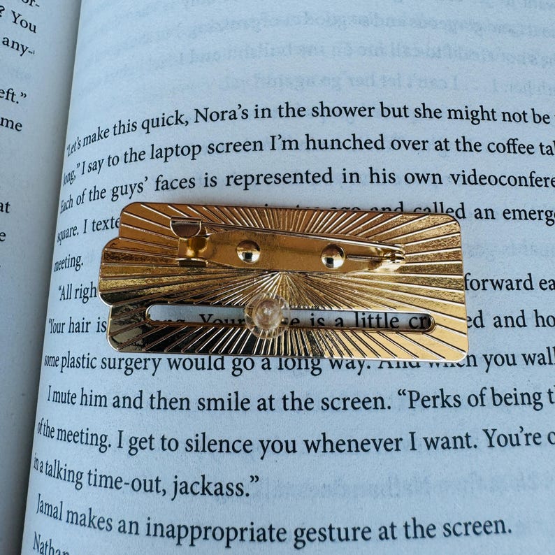 May include: A gold-colored metal pin with a sunburst design. The pin has a small, clear, round gem and the text "Your hair is a little cr". The pin is resting on the open pages of a book.