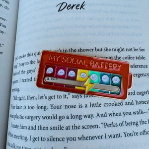 May include: A red enamel pin with the text "MY SOCIAL BATTERY" and a battery level indicator. The pin features a series of emoji faces ranging from sad to happy, and a lightning bolt. The pin is on a book page.