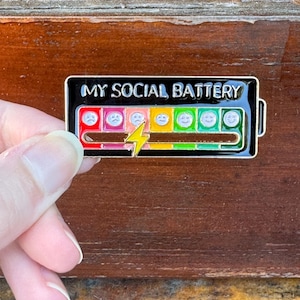May include: A black enamel pin with the text "MY SOCIAL BATTERY" at the top. The pin features a battery graphic with a range of colored emoticons, from sad to happy, and a yellow lightning bolt.