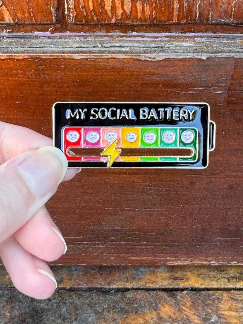 May include: A black enamel pin with the text "MY SOCIAL BATTERY" at the top. The pin features a battery graphic with a range of colored emoticons, from sad to happy, and a yellow lightning bolt.