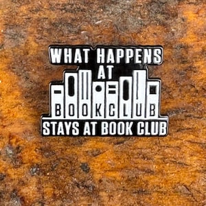 May include: A black and white enamel pin with the text "WHAT HAPPENS AT BOOK CLUB STAYS AT BOOK CLUB." The pin features a design of stylized books. The pin is set against a textured, brown background.