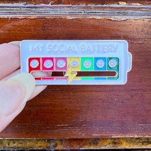 May include: A rectangular enamel pin with the text "MY SOCIAL BATTERY" at the top. The pin features a battery level indicator with a series of emoji faces ranging from sad to happy, and a lightning bolt. The pin is held by a person.