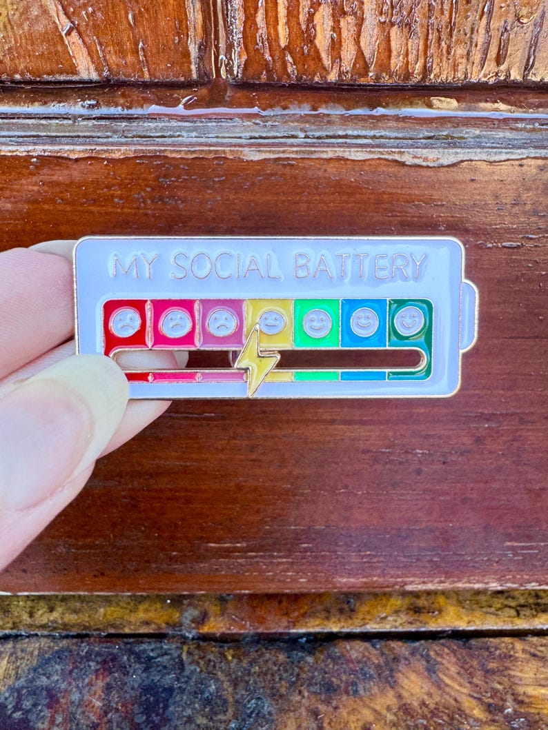 May include: A rectangular enamel pin with the text "MY SOCIAL BATTERY" at the top. The pin features a battery level indicator with a series of emoji faces ranging from sad to happy, and a lightning bolt. The pin is held by a person.
