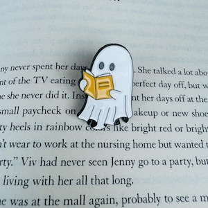 May include: A white ghost-shaped enamel pin reading a yellow book. The pin has black eyes and is set against the backdrop of an open book. The text "Fell, New York, October 1982, VIV" is visible in the background.