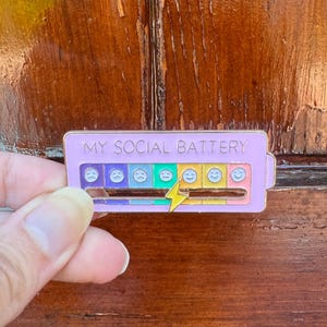 May include: A lavender enamel pin with the text "MY SOCIAL BATTERY". The pin features a battery level indicator with various emoji faces, ranging from sad to happy, and a yellow lightning bolt. The pin is rectangular in shape.