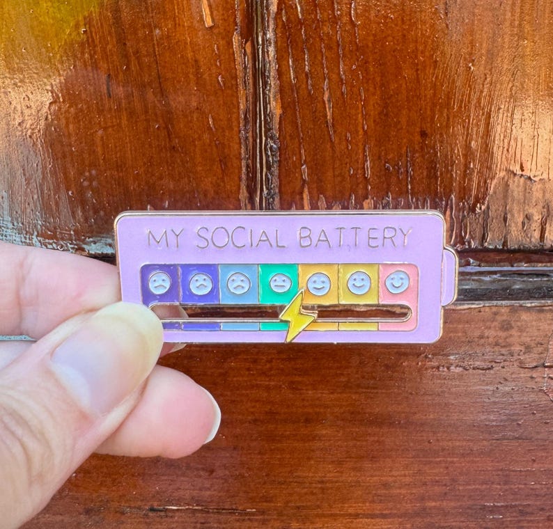 May include: A lavender enamel pin with the text "MY SOCIAL BATTERY". The pin features a battery level indicator with various emoji faces, ranging from sad to happy, and a yellow lightning bolt. The pin is rectangular in shape.